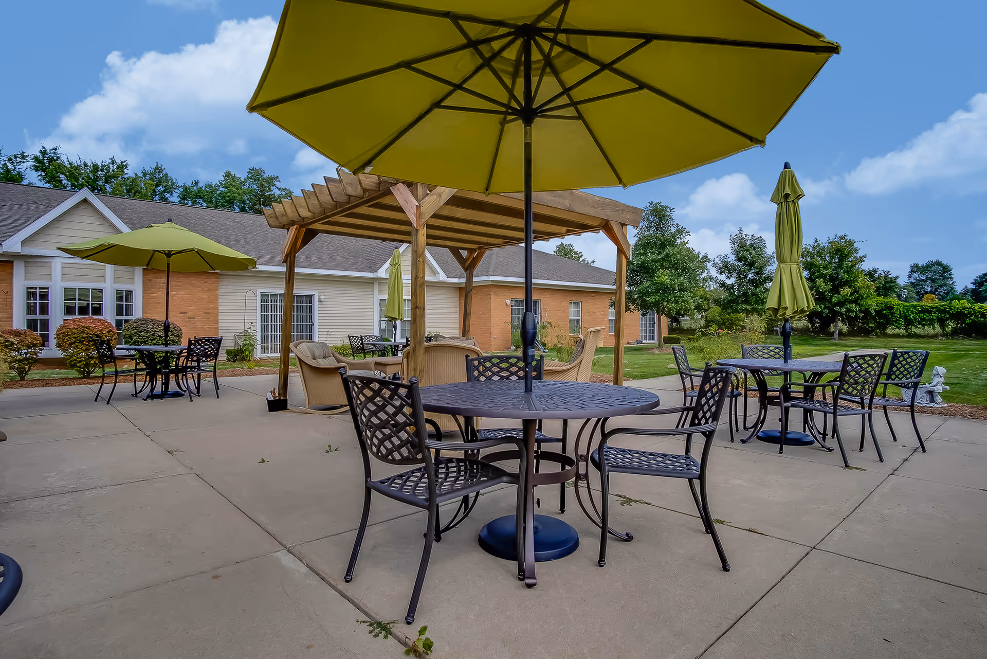 Outdoor patio with round metal tables and chairs, green umbrellas, and a wooden pergola in front of a single-story brick and siding building.