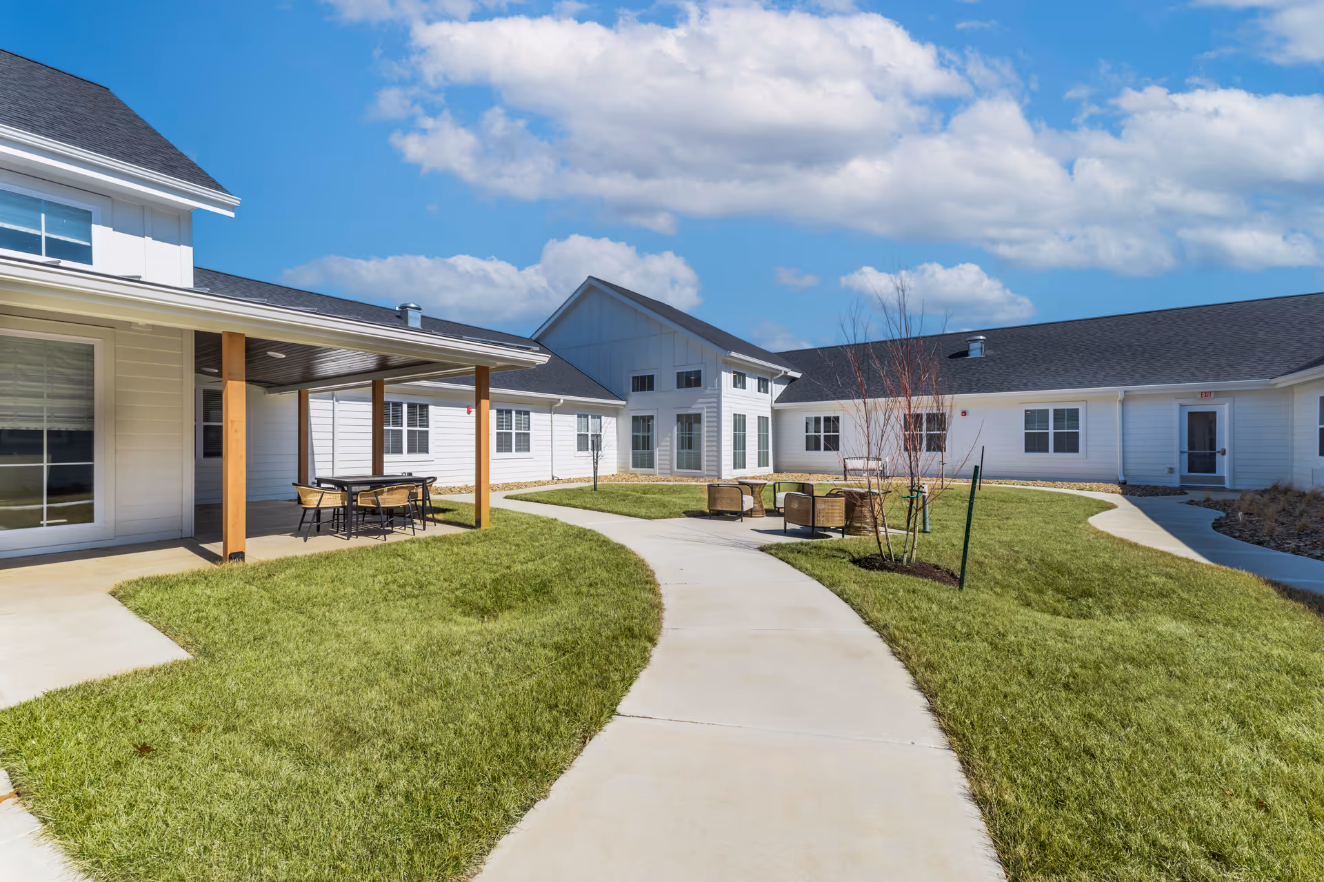Courtyard of a senior living facility with a curved concrete walkway, green lawn, covered patio with table and chairs, outdoor seating, and white one-story buildings under a blue sky.