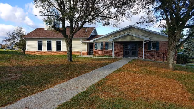 Exterior view of a single-story building with a brick and siding facade, a sloped roof, and a paved walkway leading to the entrance. There are two large trees on either side of the walkway and a grassy area surrounding the building under a partly cloudy sky.