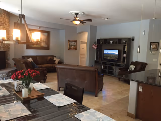 Open-plan living room and dining area with leather sofas around a TV, a dining table with place mats and flowers in the foreground, and a stone fireplace on the left.
