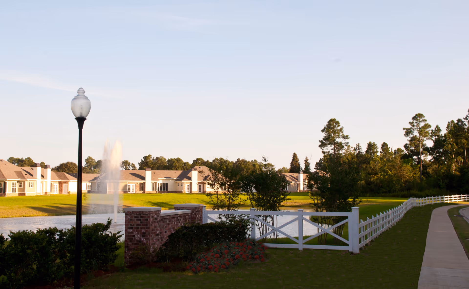 A peaceful outdoor scene at The Verandah Retirement Community featuring a paved walkway alongside a white fence, a pond with a water fountain, green lawns, bushes, and trees under a clear sky. Residential buildings are visible in the background.