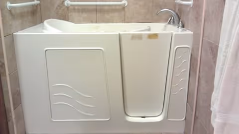 White walk-in bathtub with a side door and faucet installed against tiled walls with grab bars.