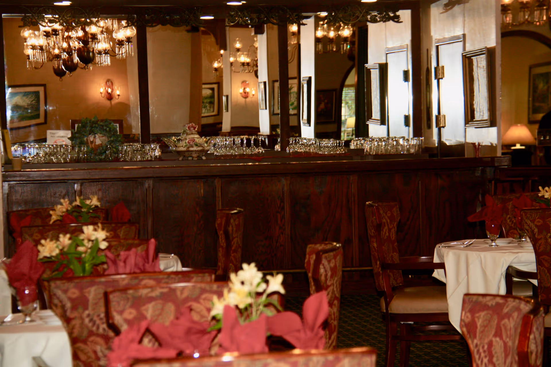 Interior view of a dining room with tables covered in white tablecloths, red cloth napkins, and floral centerpieces. The room features upholstered chairs with a patterned fabric, a large mirror on the back wall reflecting chandeliers and wall sconces, and a wooden sideboard with glassware on top.