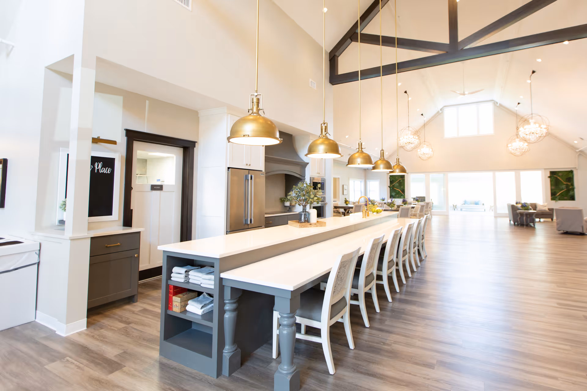 A spacious, modern dining area with a long white table and multiple white chairs lined up along it. Above the table hang six gold pendant lights. The room features high vaulted ceilings with exposed dark wooden beams and large windows letting in natural light. The floor is light wood, and there are additional seating areas with sofas in the background.
