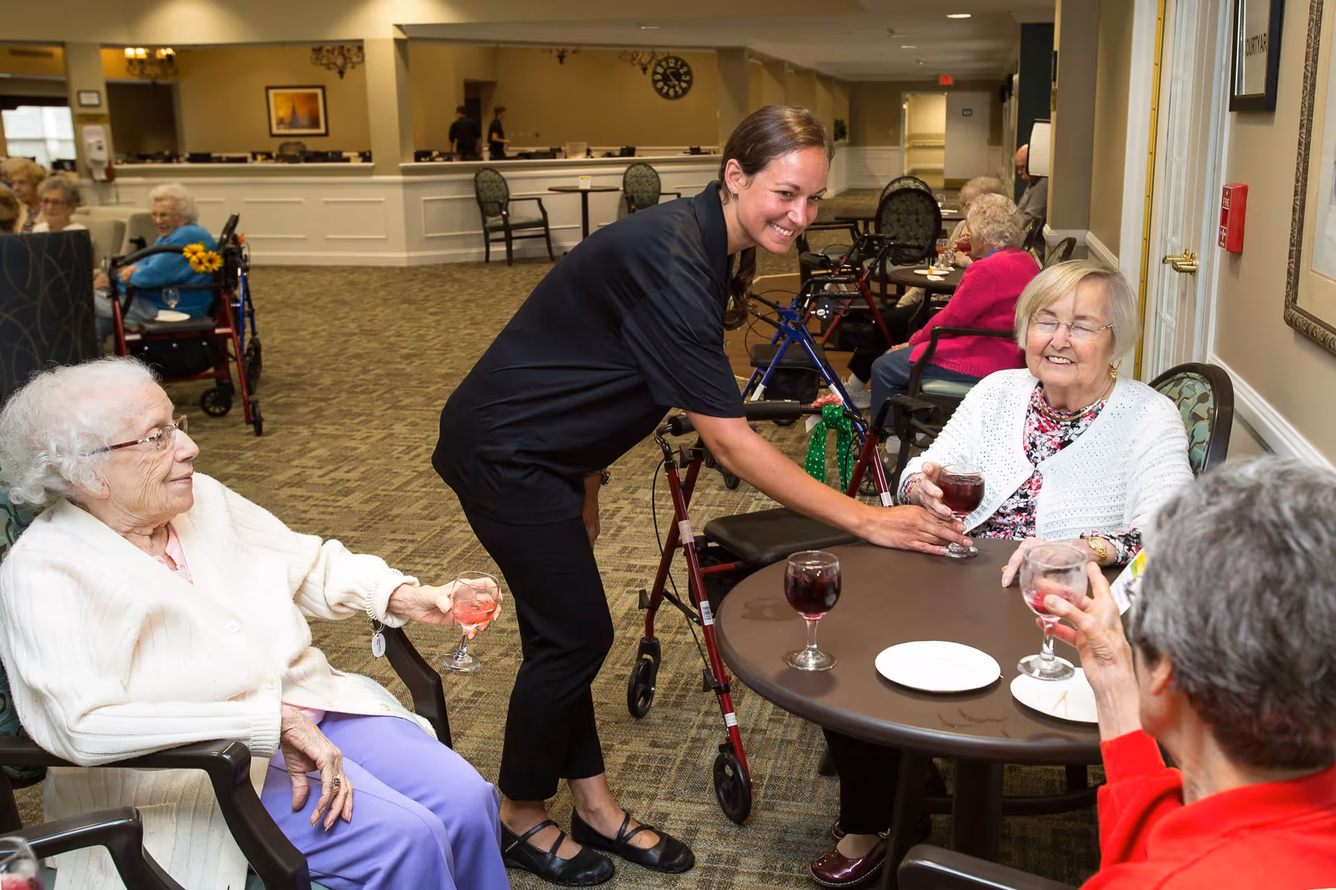 A smiling caregiver serves drinks to three elderly women seated around a round table in a communal area of a senior living facility. Other elderly residents are visible in the background, some seated in wheelchairs. The room has carpeted floors, neutral-colored walls, and comfortable chairs.