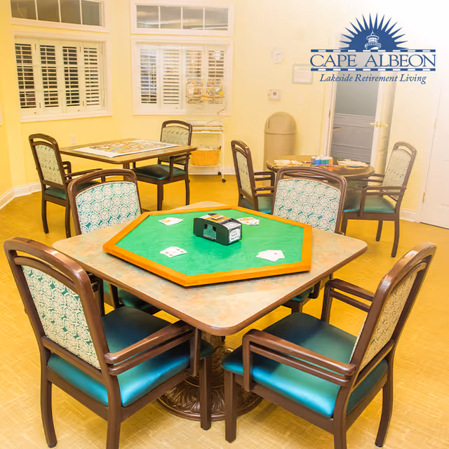 A well-lit room with multiple tables and chairs arranged for playing card games. The foreground features a square table with a green octagonal card playing surface and playing cards laid out. Other tables and chairs are visible in the background, along with windows with white shutters and a trash bin near a door. The room has a warm yellow color scheme.