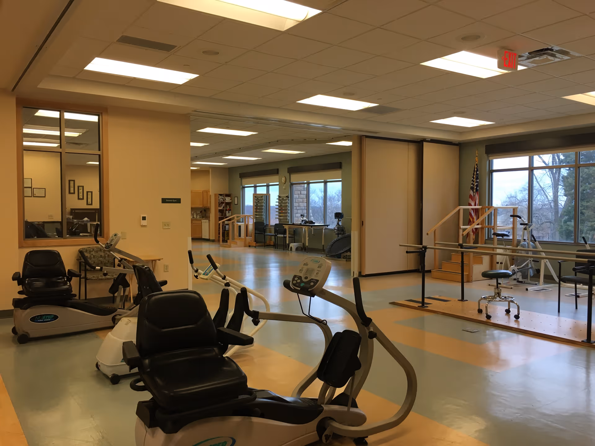 A spacious rehabilitation or physical therapy room with exercise equipment including stationary recumbent bikes and parallel bars. The room has large windows letting in natural light, an American flag in the corner, and a clean, organized environment with light-colored walls and ceiling lights.