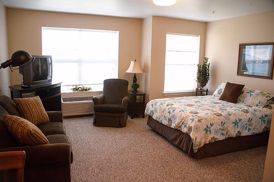 A senior living facility bedroom with a bed covered in floral bedding, a brown armchair, a sofa with striped pillows, a TV on a stand, a floor lamp, a table lamp on a side table, two windows with blinds, a framed picture on the wall, and a potted plant.