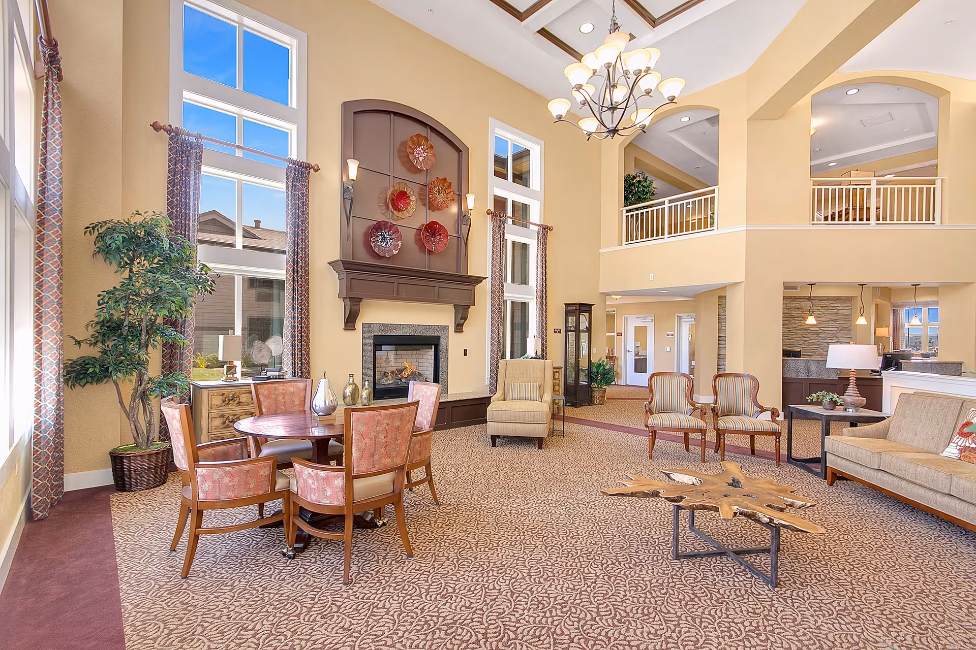 A spacious and well-lit senior living community lounge area with high ceilings and large windows. The room features a fireplace with decorative wall art above it, a round wooden table surrounded by six upholstered chairs, a beige armchair, two striped armchairs, a beige sofa, and a unique wooden coffee table. There are patterned curtains on the windows, a large potted plant, and a chandelier hanging from the ceiling. The upper level has a white railing overlooking the lounge.