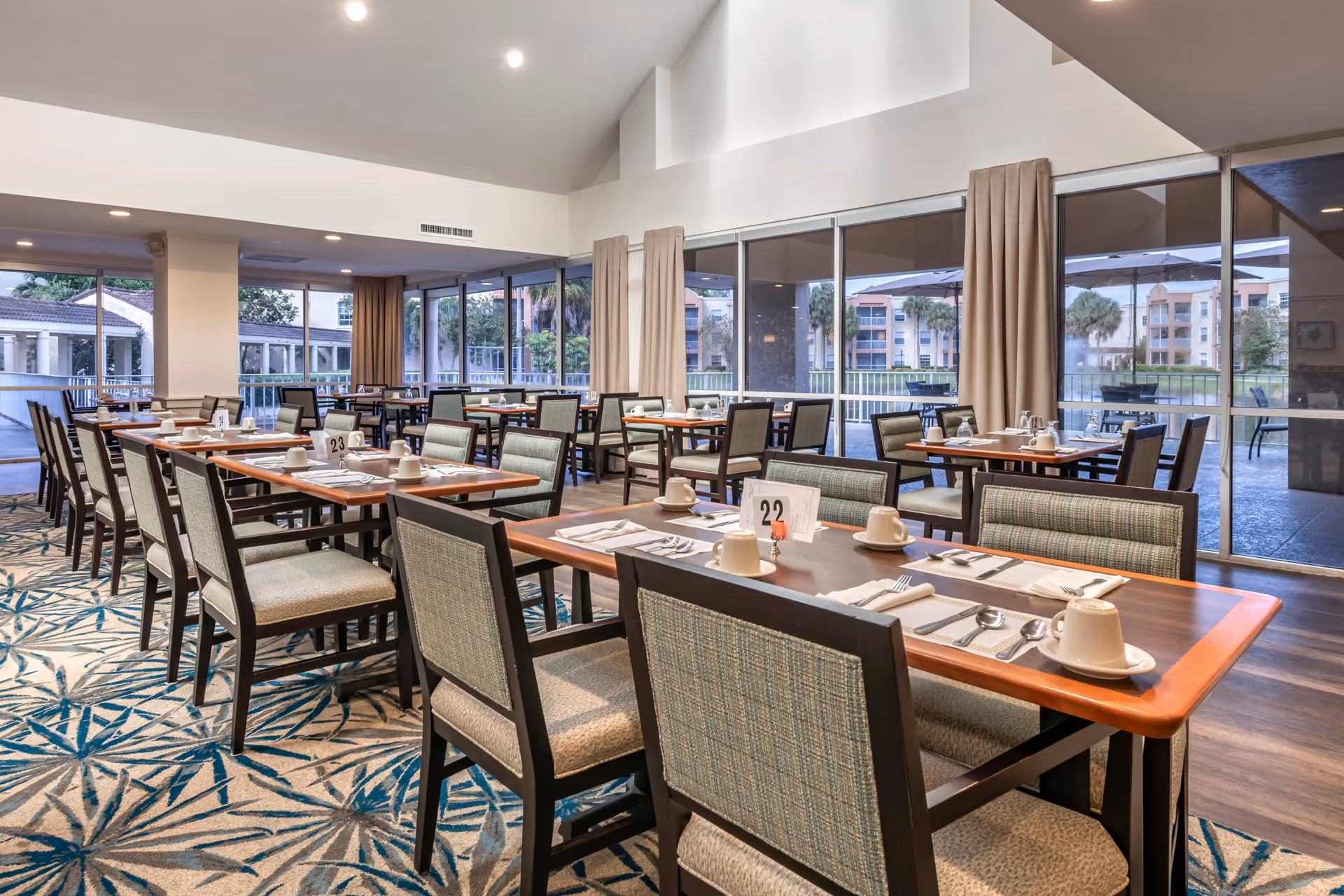 A spacious dining room with multiple tables and chairs neatly arranged. Each table is set with cups, saucers, utensils, and napkins. Large windows and glass doors provide a view of an outdoor patio area with umbrellas and greenery. The room has a high ceiling with recessed lighting and beige curtains.