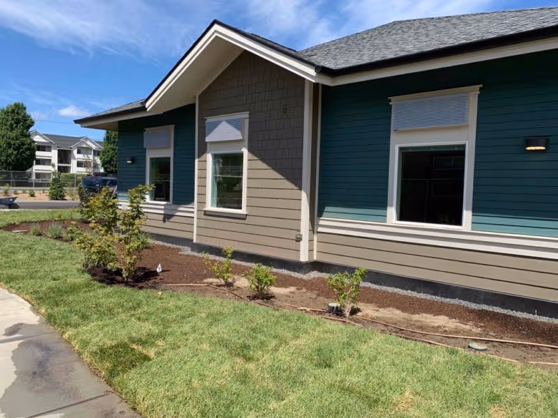 Exterior of a single-story cottage with green and beige siding, three windows, and newly landscaped grass and shrubs.