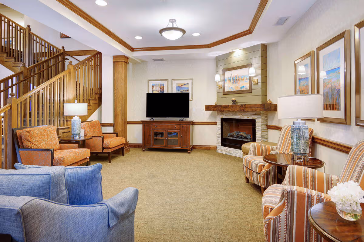 A cozy living room area in a senior living facility featuring a fireplace with a wooden mantel, a flat-screen TV on a wooden cabinet, and several upholstered chairs in orange and striped patterns. The room has beige walls with framed artwork, a carpeted floor, wooden trim, and a staircase with wooden railings in the background.