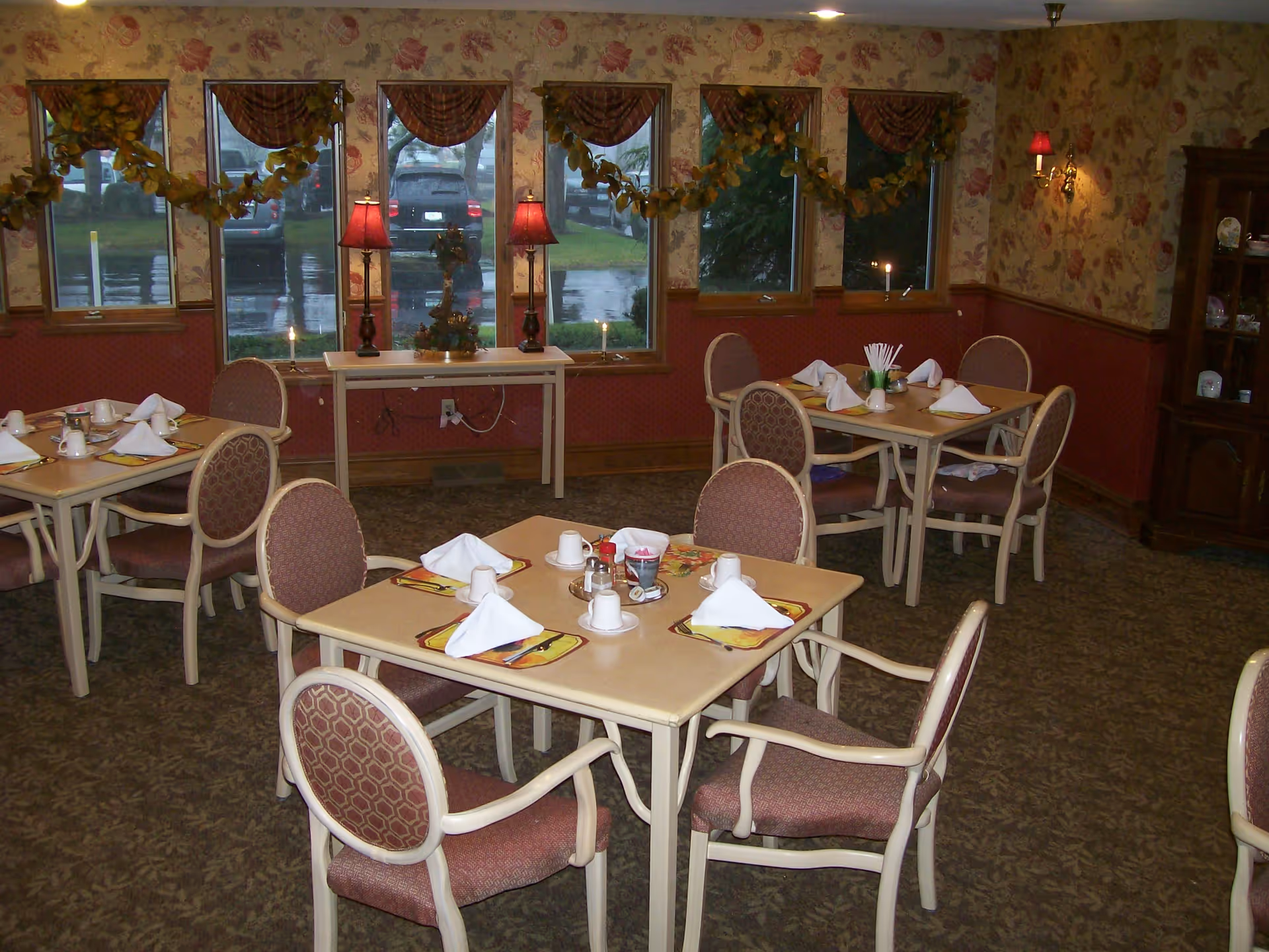 A cozy dining room with several tables set for a meal, each with placemats, napkins folded in a triangle, cups, and condiments. The room has floral wallpaper, carpeted floor, and windows with valances. There are two red lamps on a small table against the wall and a garland decoration hanging across the windows. A wooden cabinet with dishes is visible in the corner.