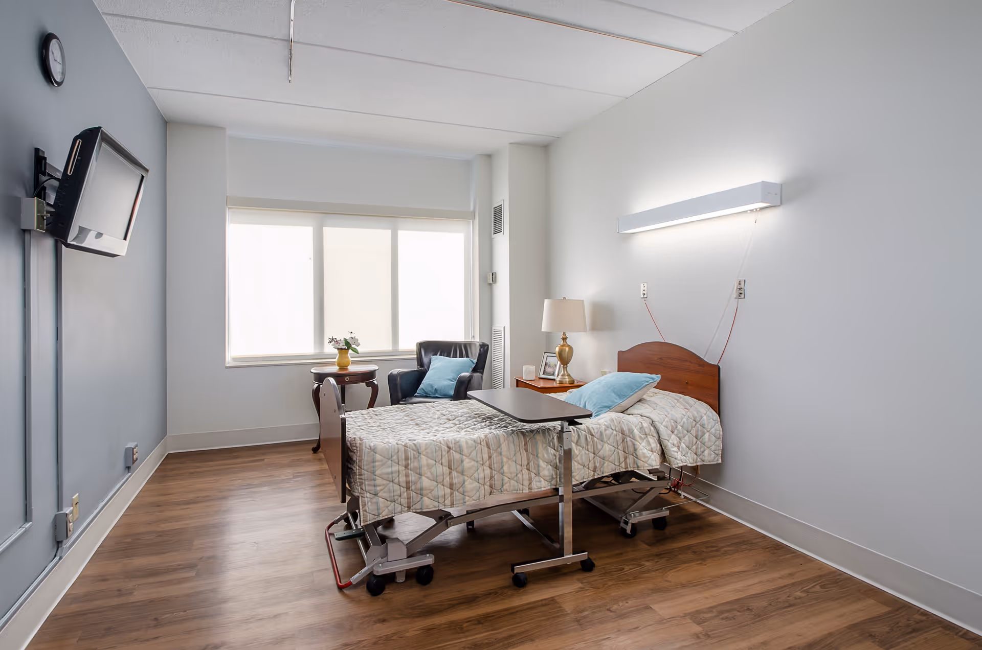 A simple, clean bedroom in a senior living facility with a hospital-style bed covered with a patterned quilt, a small overbed table, a wooden nightstand with a lamp and picture frames, a black armchair with a blue pillow, a small round table with a vase of flowers, a wall-mounted TV, and a large window letting in natural light.
