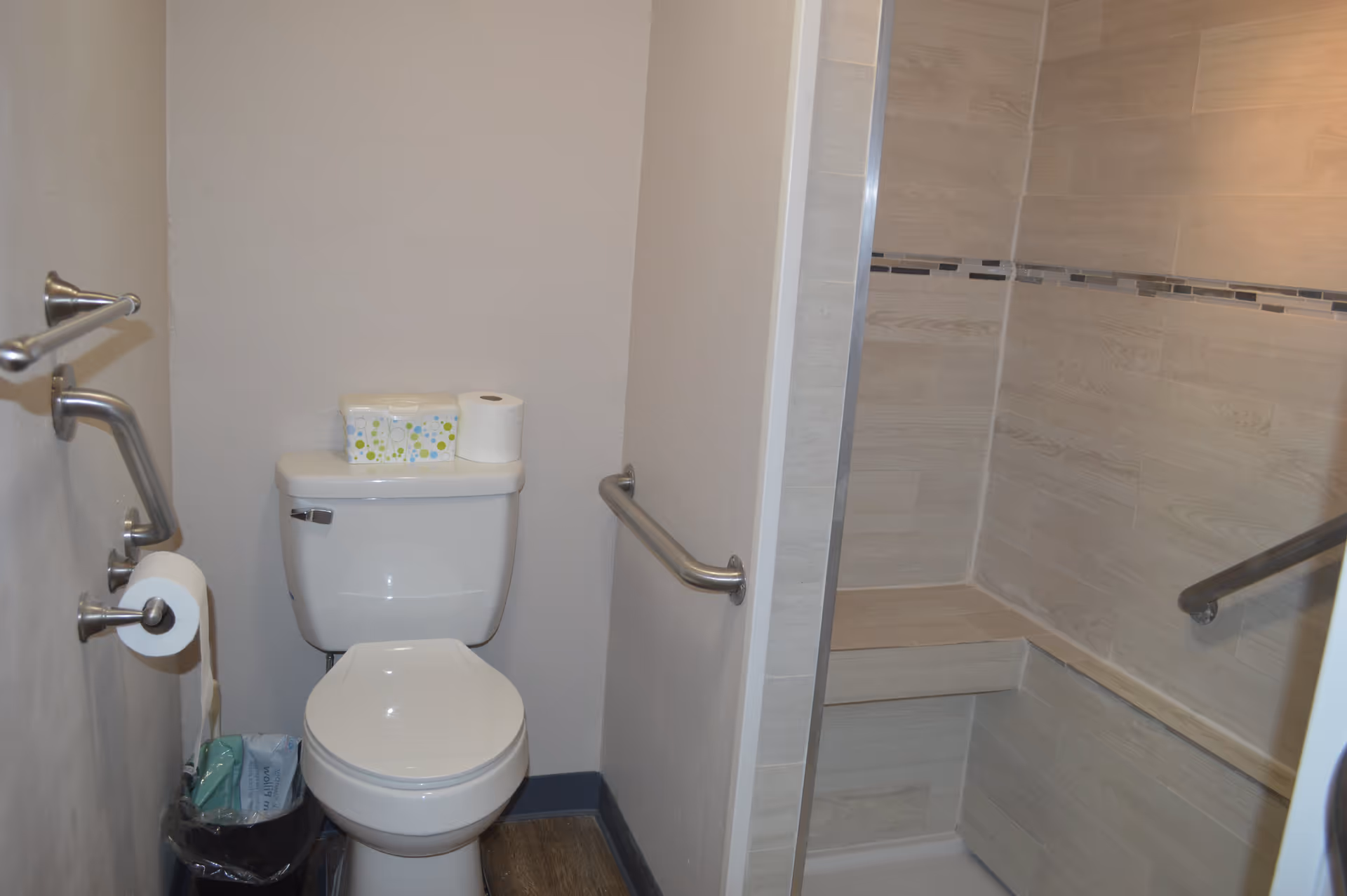 A bathroom with a white toilet, a roll of toilet paper on a holder, a box of wipes and an extra roll of toilet paper on top of the toilet tank. The bathroom has grab bars on the walls and a tiled shower area with a built-in bench and grab bar.