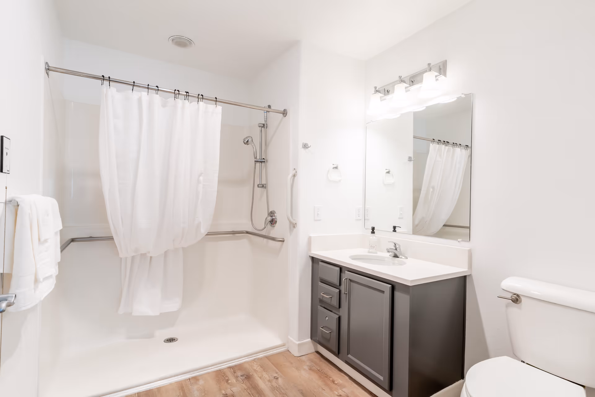 A clean and modern bathroom featuring a walk-in shower with a white curtain, a gray vanity with a white countertop and sink, a large mirror with three light fixtures above it, a toilet, and wooden flooring.