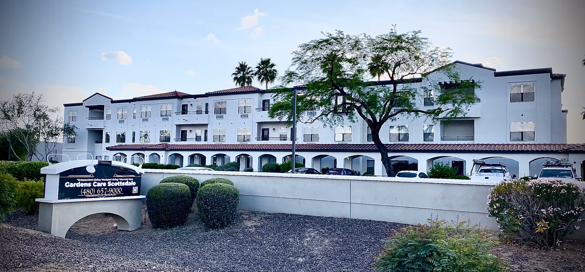 Exterior view of a multi-story senior living facility building with white walls and red-tiled roof accents. The foreground features a landscaped area with bushes and a sign that reads 'Gardens Care Scottsdale' along with a phone number. Several cars are parked near the building under a covered area.
