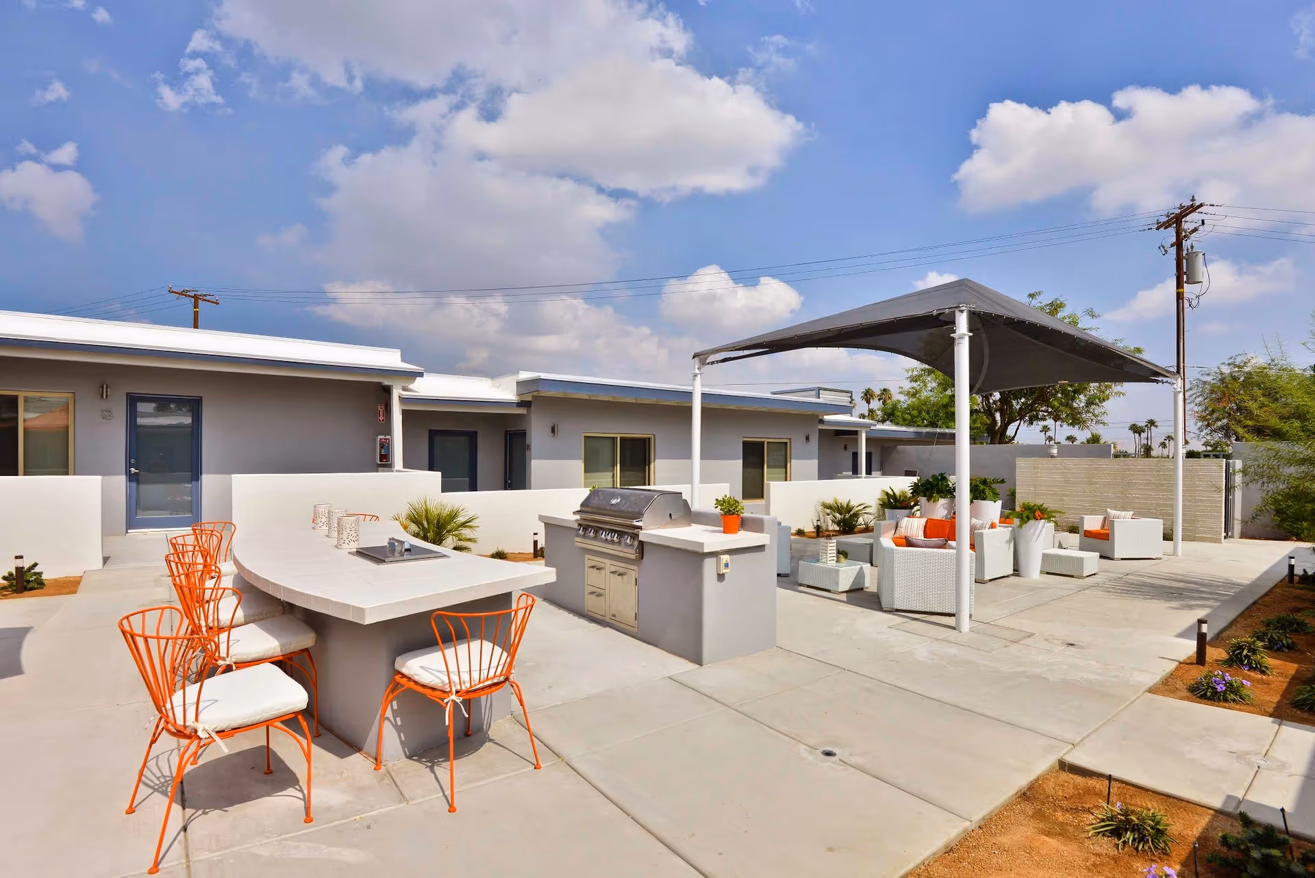 Outdoor patio area at Stonewall Gardens Assisted Living featuring a built-in grill, a curved bar counter with orange metal chairs, and a shaded seating area with white wicker furniture and orange cushions under a canopy. The setting is bright with a partly cloudy sky and surrounding low buildings.