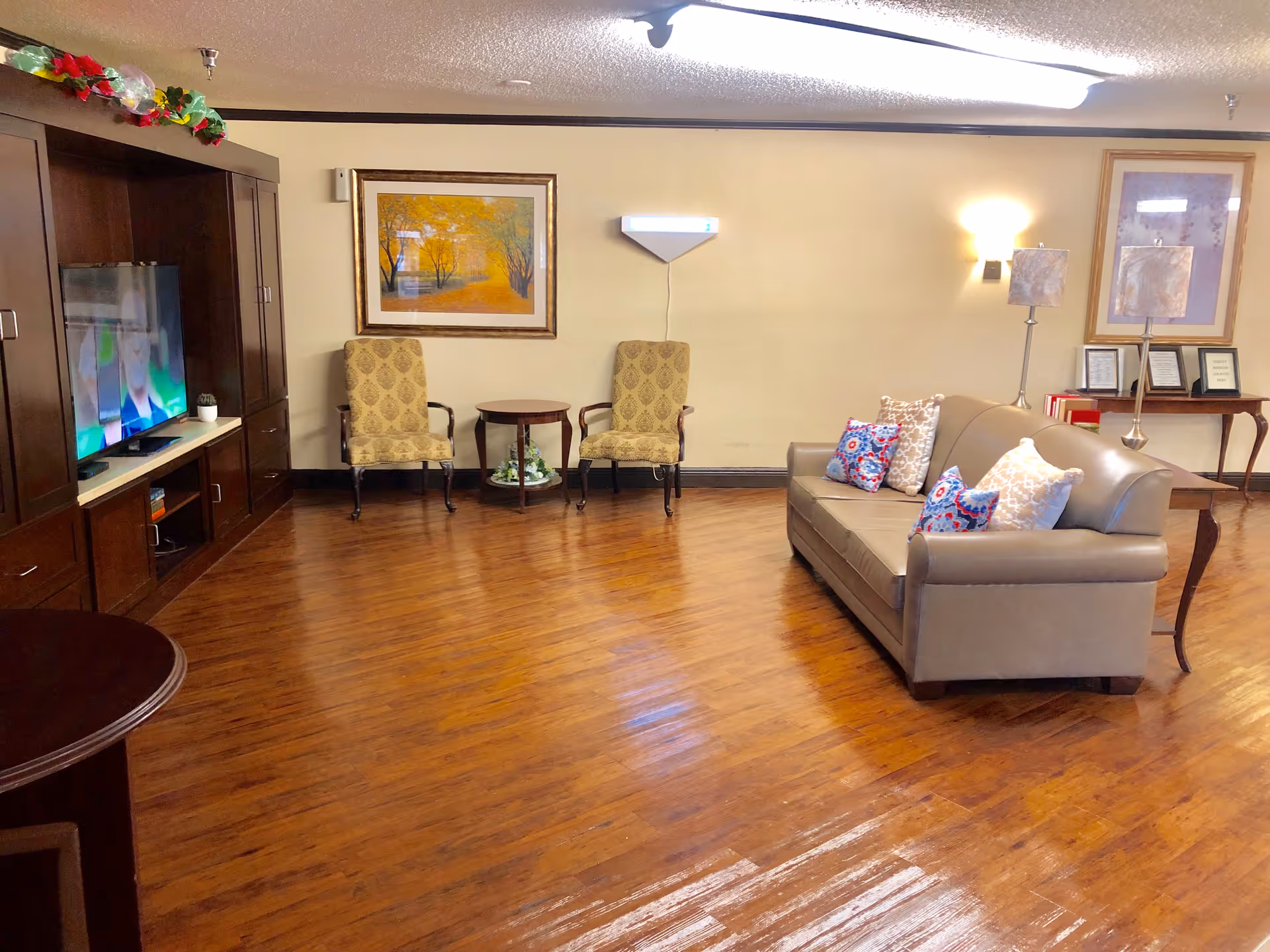 A cozy living room area with wooden flooring, a beige leather sofa adorned with colorful patterned pillows, two upholstered armchairs with a small round table between them, a large wooden entertainment center with a TV, framed artwork on the walls, and soft lighting from wall sconces and lamps.