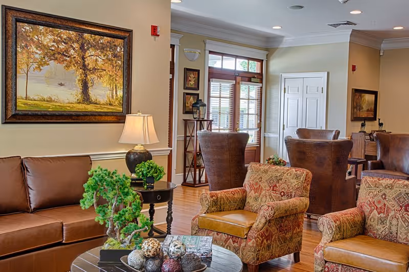 A cozy senior living facility common area with a brown leather sofa, patterned armchairs, a round coffee table with decorative items, a side table with a lamp and plant, wooden flooring, and framed artwork on the walls. There is a door with glass panels and windows letting in natural light.
