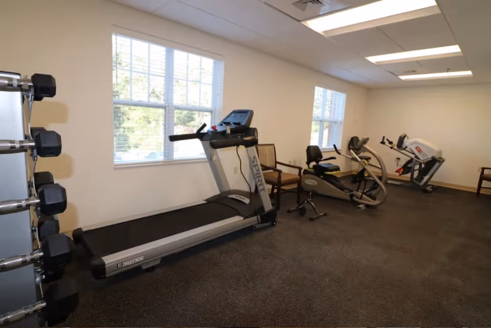 A small fitness room with exercise equipment including a treadmill, a recumbent bike, and a weight machine. There are two windows letting in natural light, a rack of dumbbells on the left, and a couple of chairs placed near the equipment.
