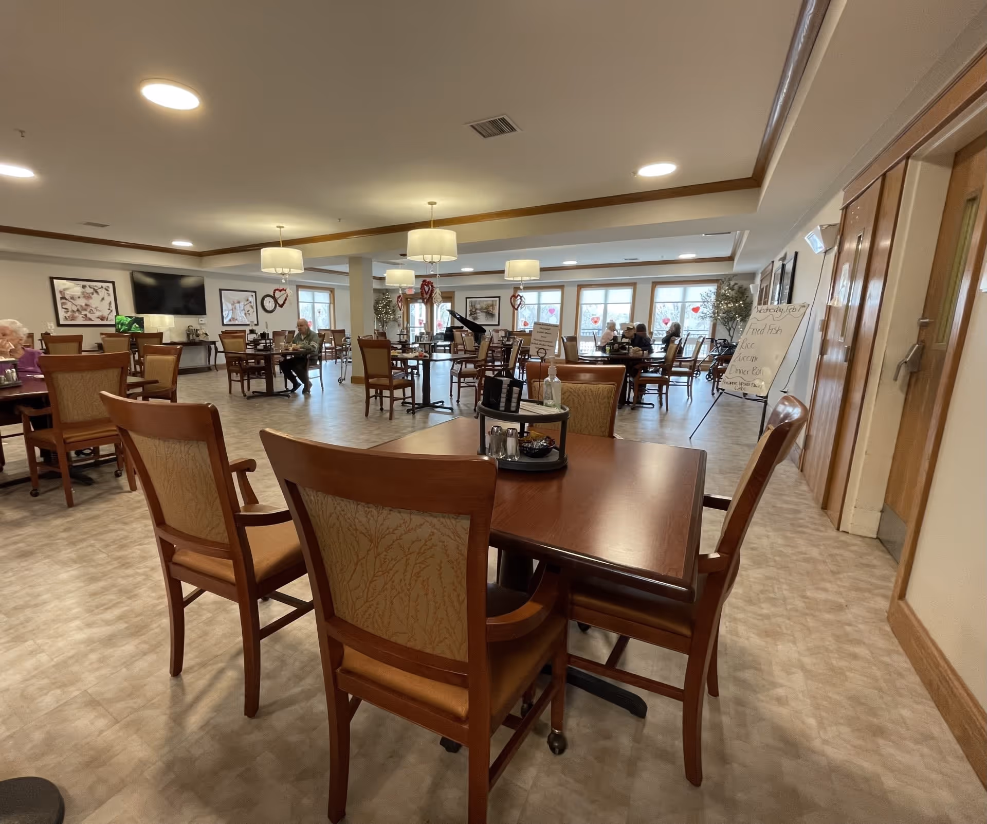 A spacious dining room in a senior living facility with multiple wooden tables and chairs. Several elderly residents are seated at the tables. The room is well-lit with ceiling lights and large windows decorated with heart-shaped decals. There is a piano in the background and framed artwork on the walls.