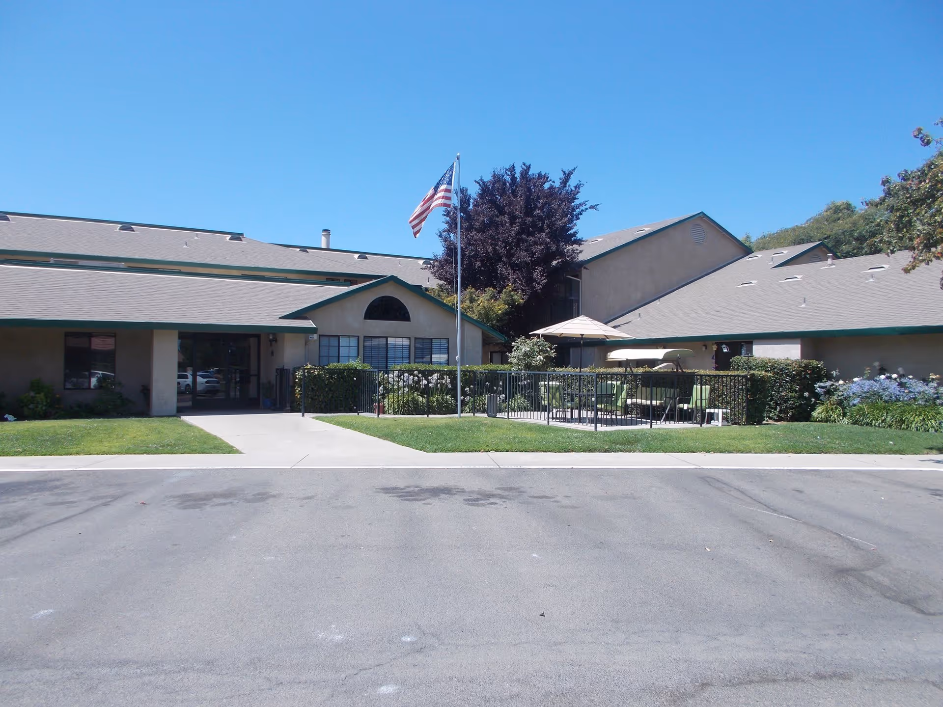 Exterior view of a single-story building with a gray roof and beige walls, featuring a small fenced patio area with outdoor furniture and an umbrella. An American flag is flying on a flagpole in front of the building, with a clear blue sky overhead.