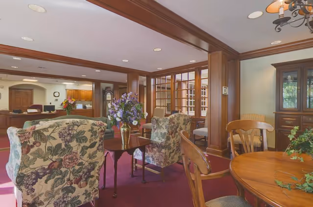 A cozy and well-lit common area in Glen Meadows Retirement Community featuring floral upholstered armchairs around a wooden coffee table with a vase of purple flowers. The room has wooden paneling, a grandfather clock, and a wooden cabinet. There is a round wooden table with chairs in the foreground and a reception desk in the background.