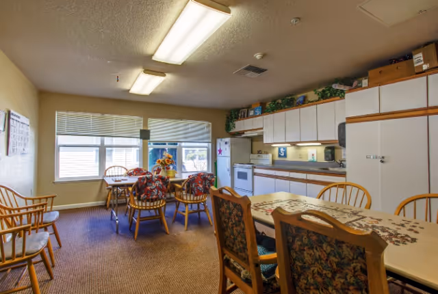 Communal dining area with several tables and wooden chairs, floral cushions, and a kitchenette with white cabinets and appliances under fluorescent lights.