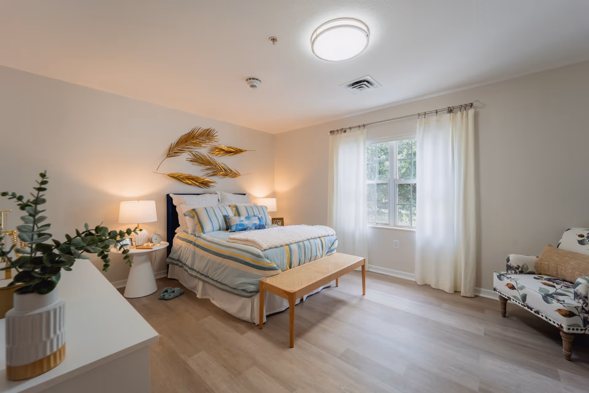 A bright and cozy bedroom with a neatly made bed featuring striped bedding and multiple pillows. There are two bedside tables with lamps on either side of the bed. Above the bed, there is a decorative wall art piece with golden leaves. A window with white curtains allows natural light to fill the room. A floral-patterned armchair with a cushion is placed near the window, and a wooden bench is at the foot of the bed. A white dresser with a plant in a white and gold vase is visible in the foreground.