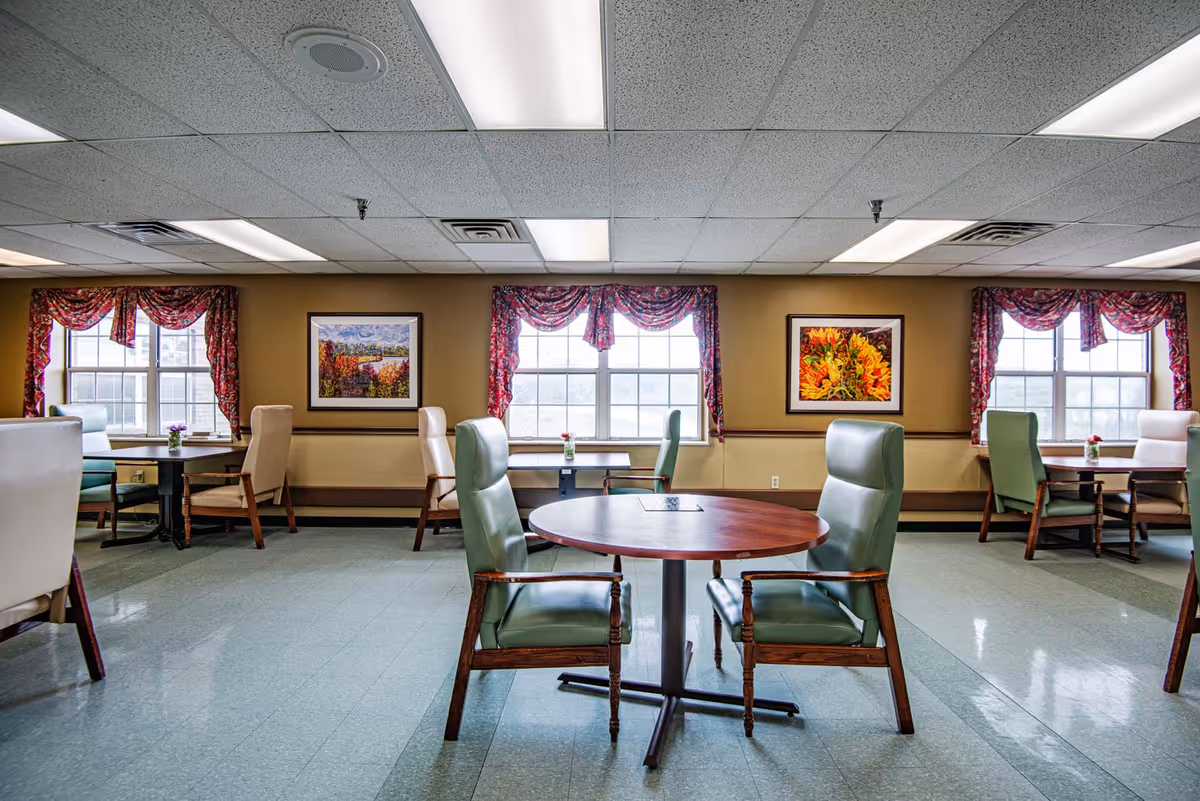 A bright dining room with multiple round and rectangular tables surrounded by cushioned chairs. The room has large windows with red patterned valances, framed artwork on the walls, and a tiled floor. The ceiling has recessed lighting panels.