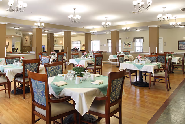 A spacious dining room with multiple round tables covered with white and green tablecloths, each set with napkins, glasses, and small flower arrangements. The room has wooden floors, several chandeliers hanging from the ceiling, and large windows letting in natural light.