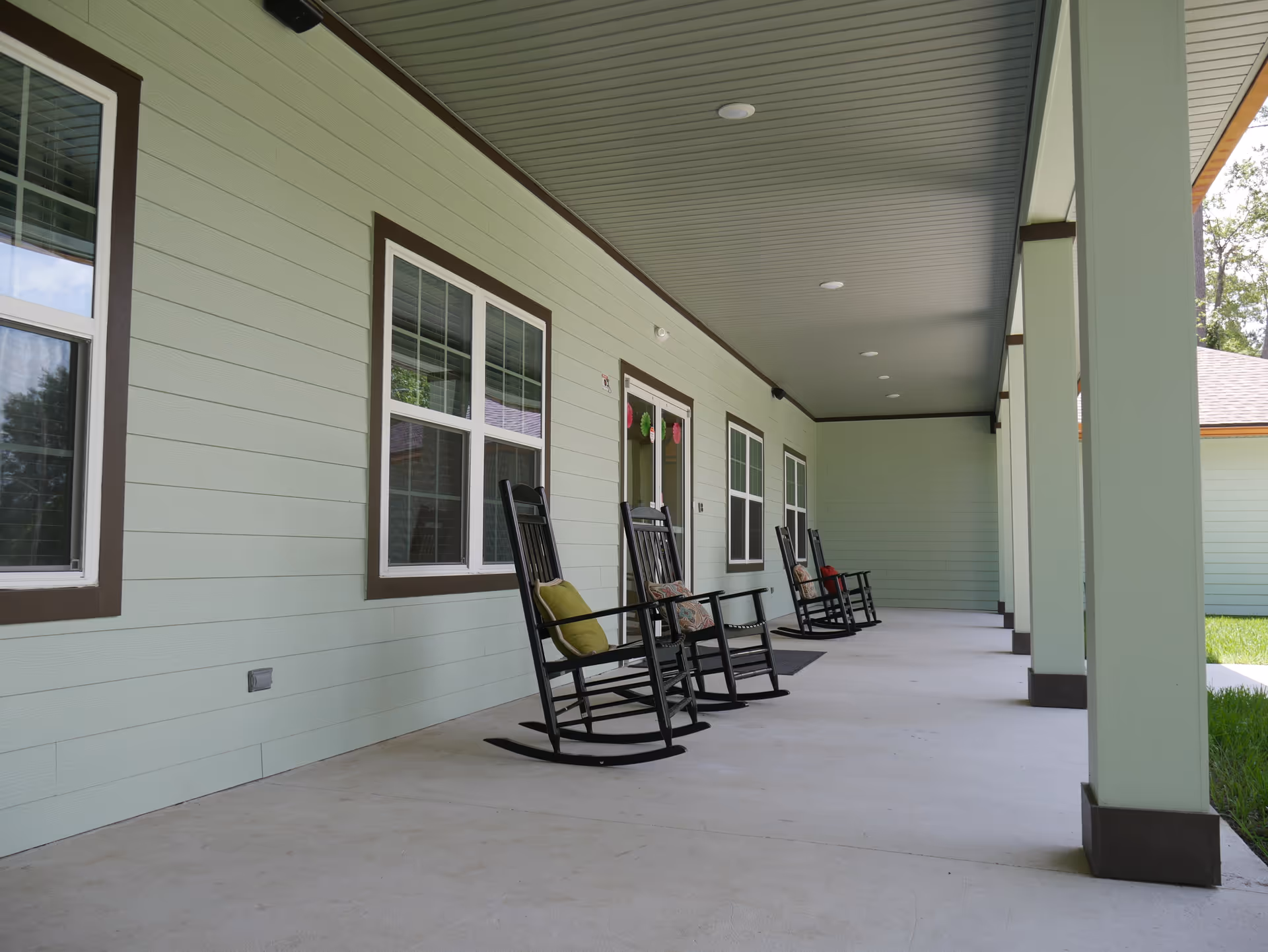 A covered outdoor porch area with light green walls and ceiling, featuring several black rocking chairs with cushions lined up along the wall under recessed lighting. There are multiple windows and a door decorated with hanging ornaments, and green grass is visible beyond the porch.