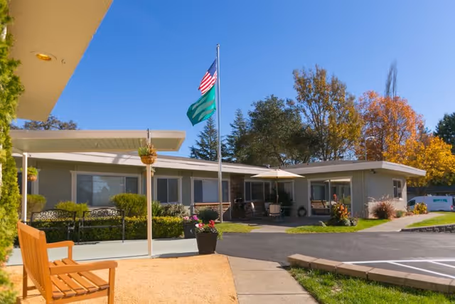 Exterior view of Summerfield Healthcare Center showing a single-story building with large windows, a flagpole with the American flag and another green flag, a wooden bench, hanging flower pots, and a paved walkway surrounded by greenery and trees with autumn foliage under a clear blue sky.