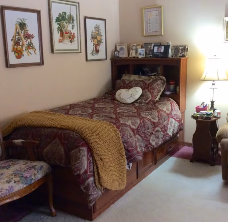 Cozy single bedroom with a patterned bed and wooden headboard topped with framed photos, a lamp on a side table, and floral wall art on the pale walls.