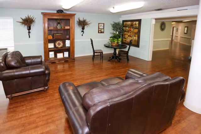 A lounge area with leather sofas, a bookshelf, a small table and chairs, and hardwood floors in a senior living facility.