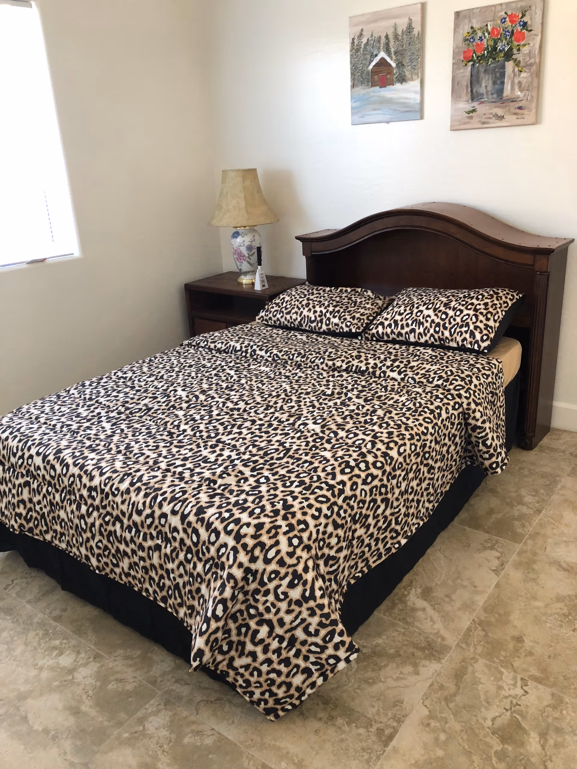 A bedroom with a bed covered in leopard print bedding and two matching pillows. There is a wooden headboard and a nightstand with a floral lamp. Two paintings hang on the wall above the bed, one depicting a snowy cabin and the other a bouquet of flowers. A window with blinds is partially visible on the left side.