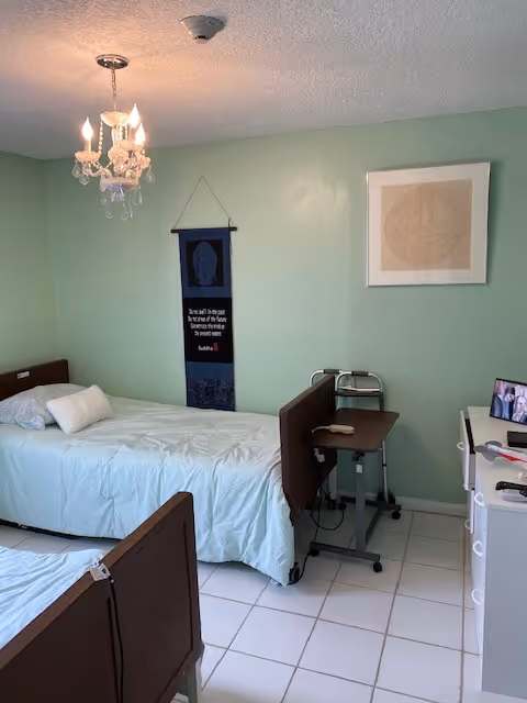 A small, simple bedroom with two single beds with wooden headboards and footboards. One bed is fully visible with a white pillow and light blue bedding. A small overbed table with a remote control is positioned next to the bed. The walls are painted light green and decorated with a hanging tapestry and a framed abstract artwork. A chandelier-style light fixture hangs from the ceiling. A white dresser with a few items on top is visible on the right side of the image.