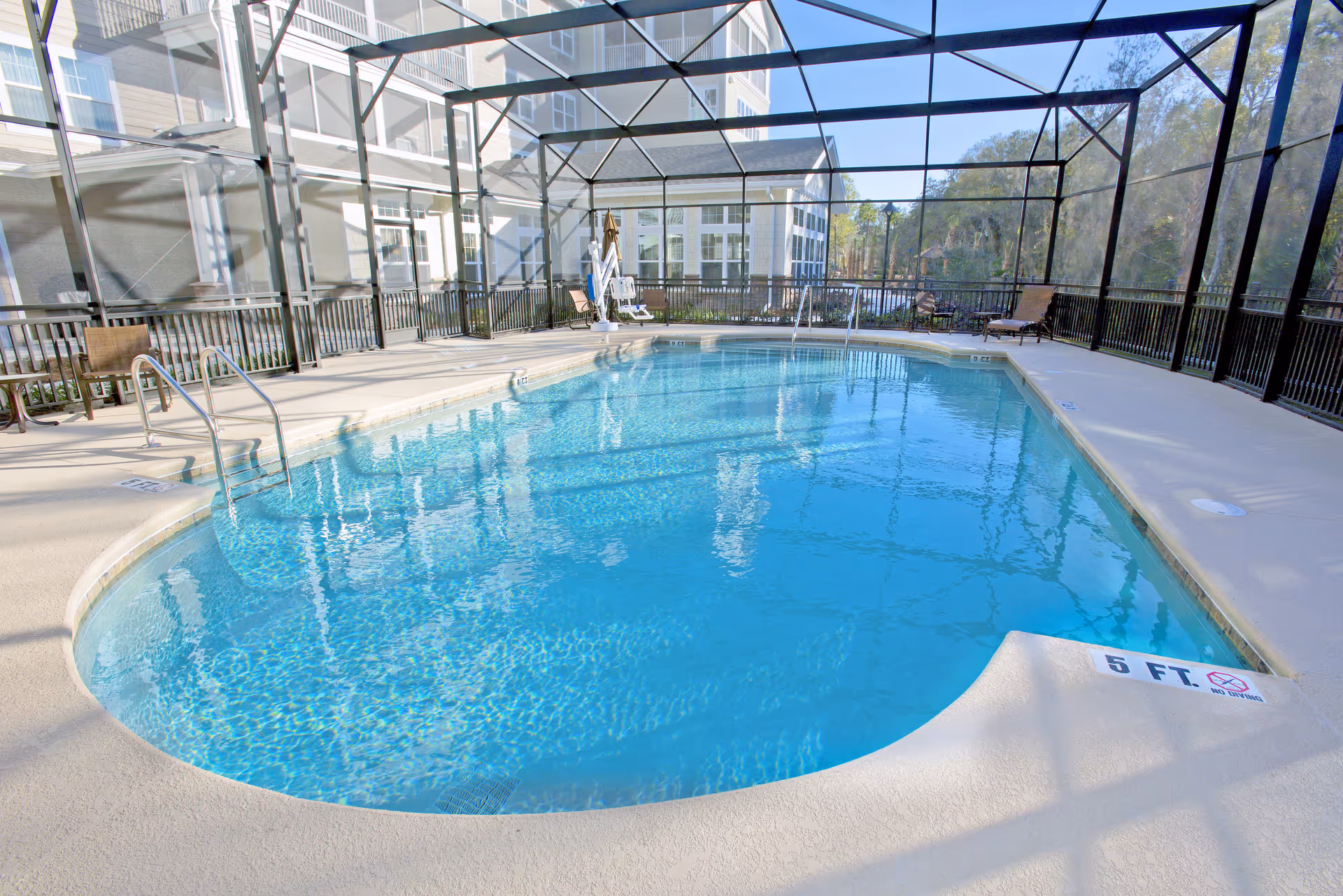 Indoor swimming pool with clear blue water enclosed by a screened structure. The pool area has a safety railing, pool lift, and several lounge chairs around the perimeter. The building exterior and trees are visible through the screened enclosure.