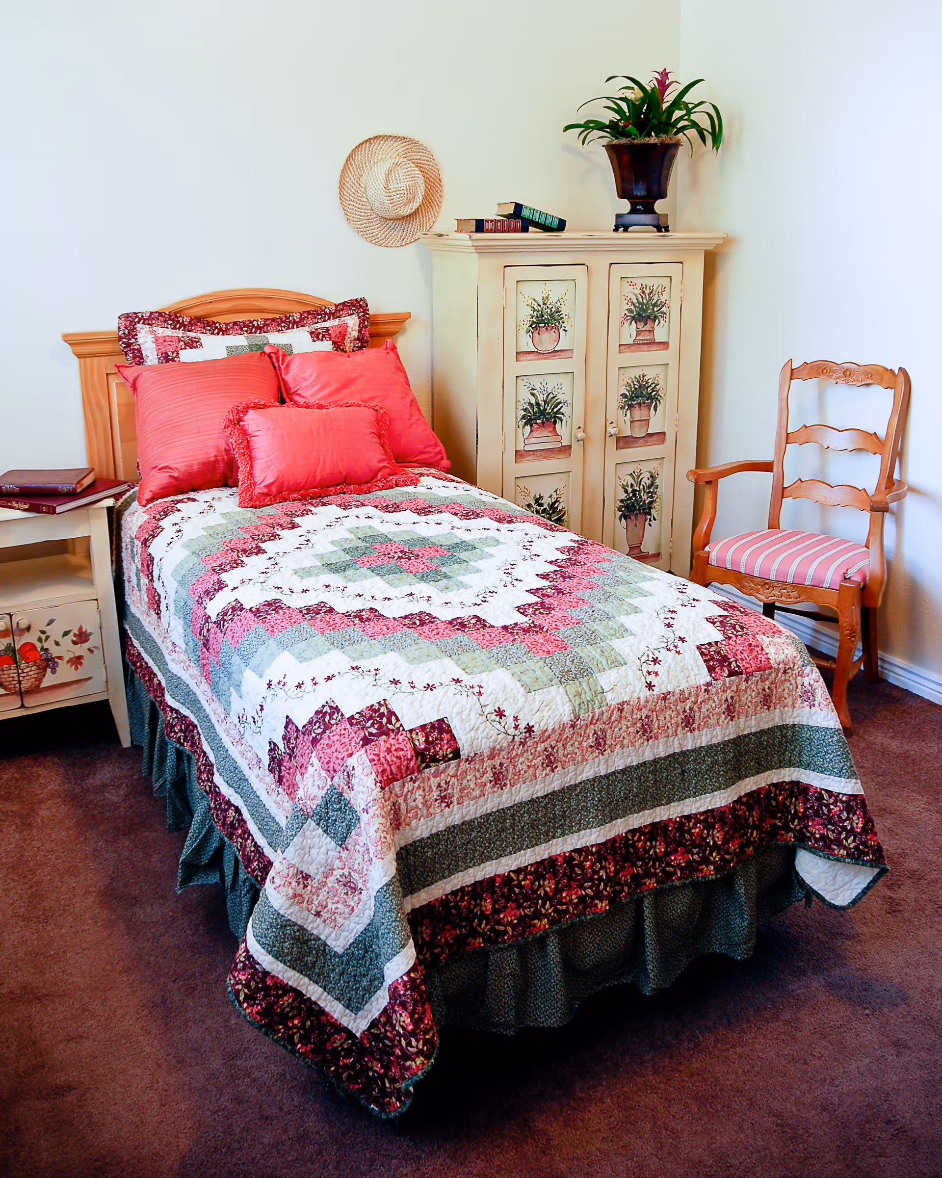 A cozy bedroom with a single bed covered in a colorful patchwork quilt featuring shades of red, green, and white. The bed has several red pillows and a wooden headboard. Next to the bed is a small nightstand with books on it. A decorative cabinet with floral paintings and a potted plant on top is against the wall. A wooden chair with a pink striped cushion is placed beside the cabinet. A woven hat hangs on the wall above the bed.