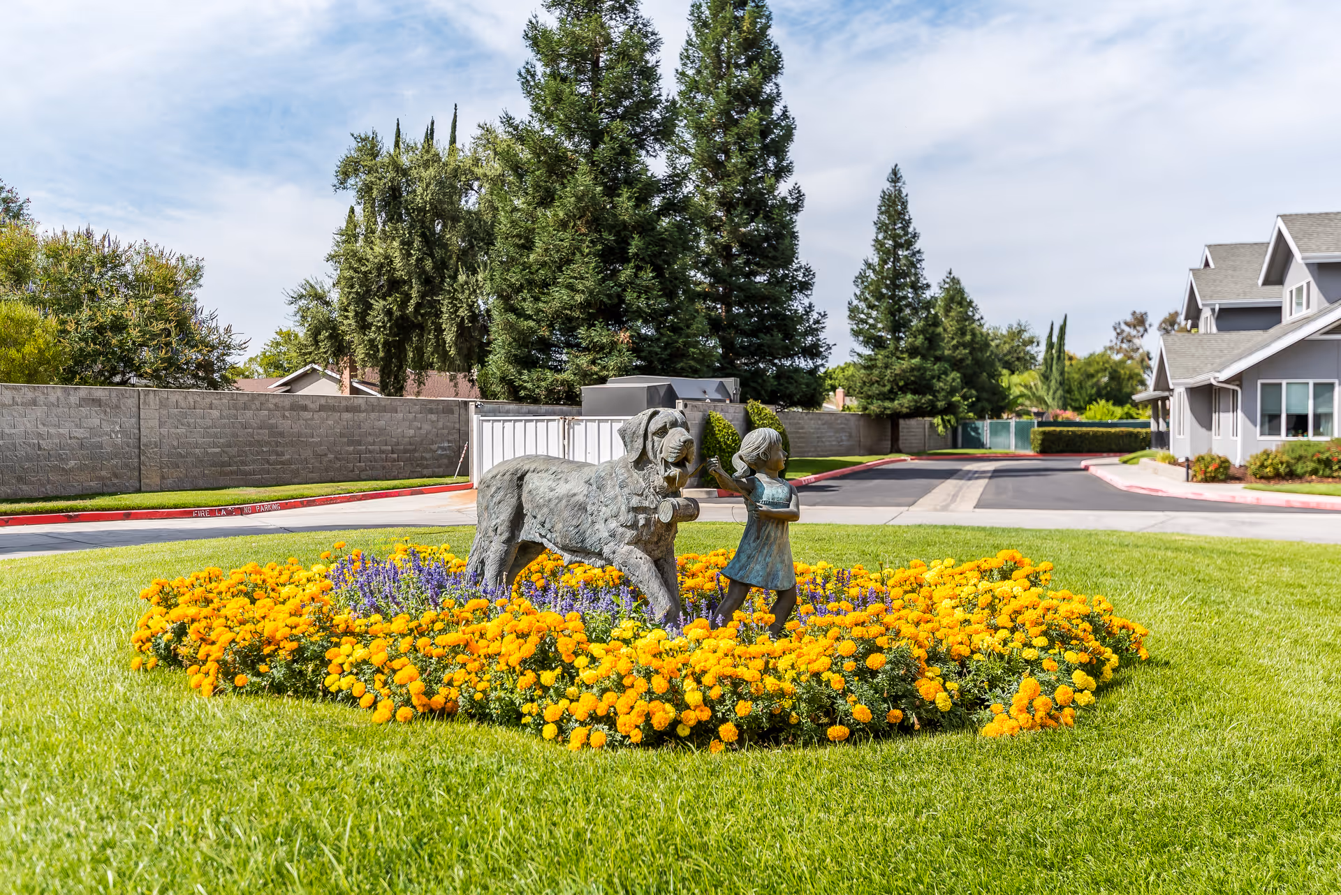A landscaped outdoor area with a circular flower bed filled with yellow and purple flowers. In the center of the flower bed is a bronze statue of a girl and a dog. The background shows a paved road, a stone wall, tall trees, and residential-style buildings under a partly cloudy sky.