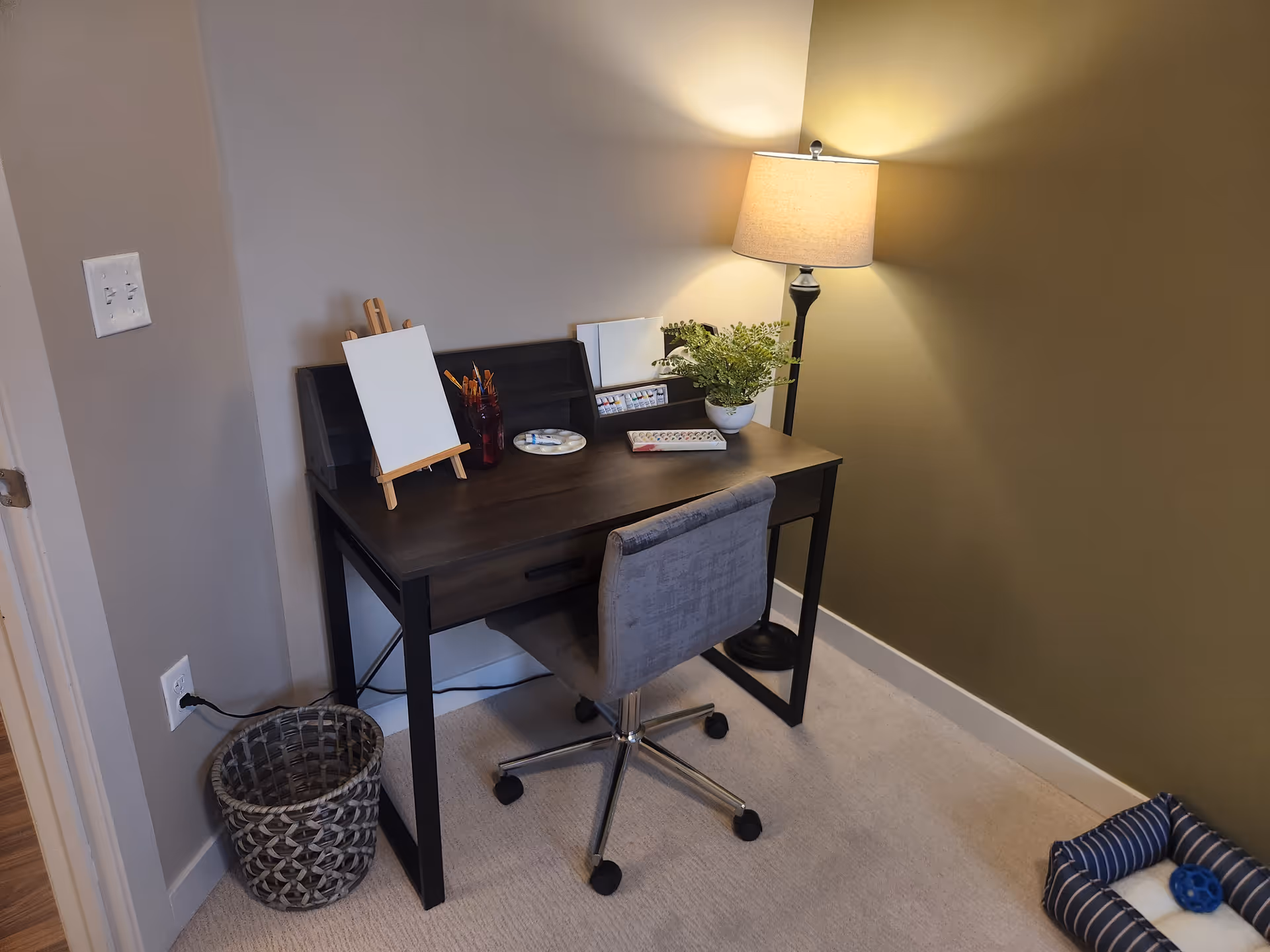 Small interior workspace with a dark wood desk, gray rolling chair, table lamp, potted plant and art supplies in a corner.