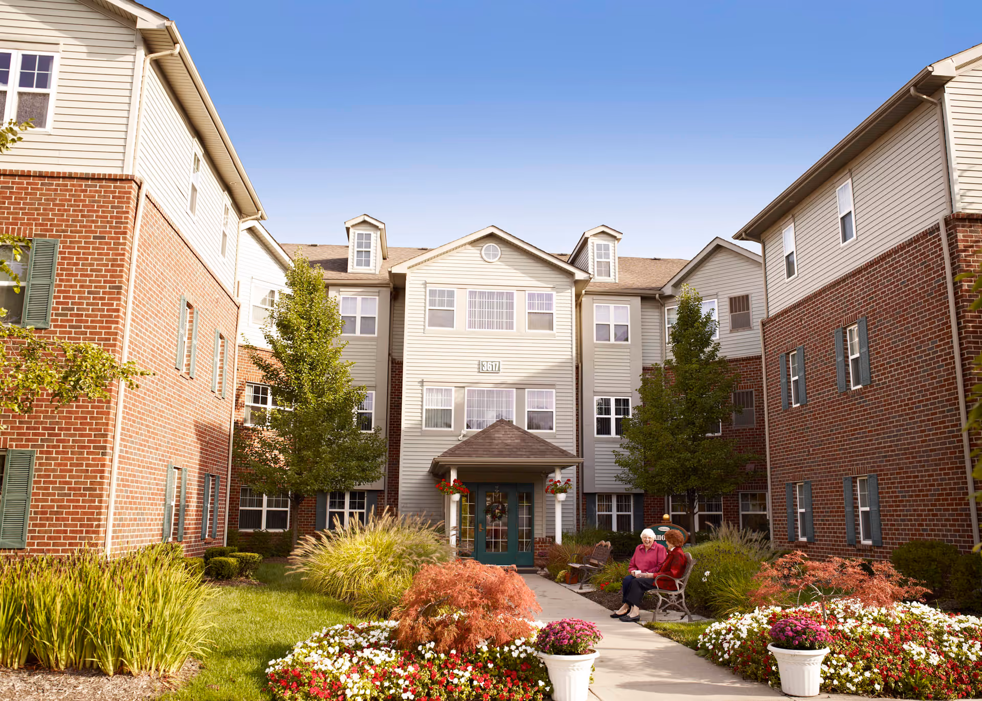 Exterior view of a senior living facility with a well-maintained garden and walkway leading to the entrance. Two elderly women are sitting and chatting on benches near the entrance, surrounded by colorful flowers and greenery. The building features a combination of brick and siding with multiple windows and dormer windows on the roof under a clear blue sky.