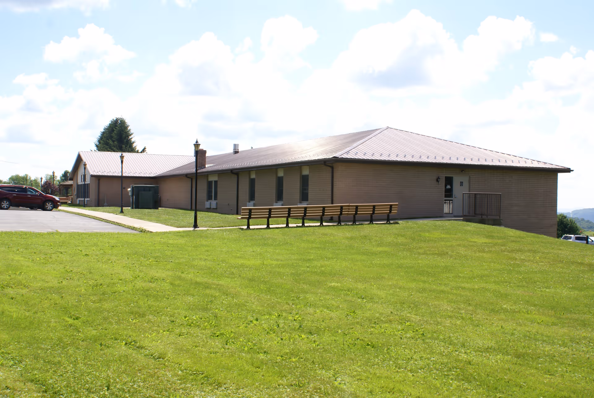 Exterior view of a single-story brick building with a metal roof, surrounded by a well-maintained grassy lawn. There are several benches lined up on the grass near the building, a sidewalk leading to a parking lot with a red car, and lamp posts along the walkway under a partly cloudy sky.