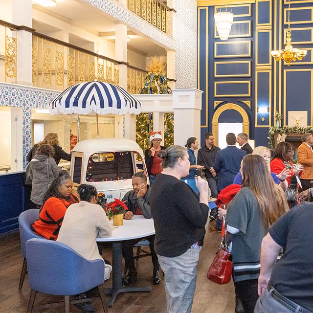Festively decorated lobby with people gathered around tables and a small striped-canopy cart in the interior common area.