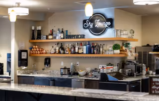 Interior view of a bar area named Sky Lounge with shelves holding various bottles of alcohol, glasses, and decorative items. The bar counter is visible in the foreground with a granite countertop, and there are pendant lights hanging from the ceiling.
