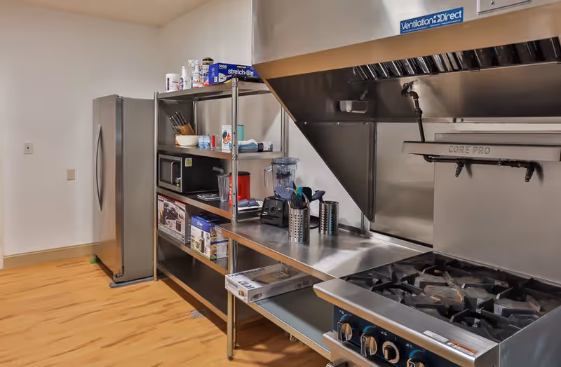 A commercial kitchen area with a stainless steel refrigerator, metal shelving holding various kitchen items including a microwave, blender, and kitchen utensils. There is a large stainless steel stove with multiple burners and an industrial ventilation hood above it. The floor is wooden and the walls are plain white.