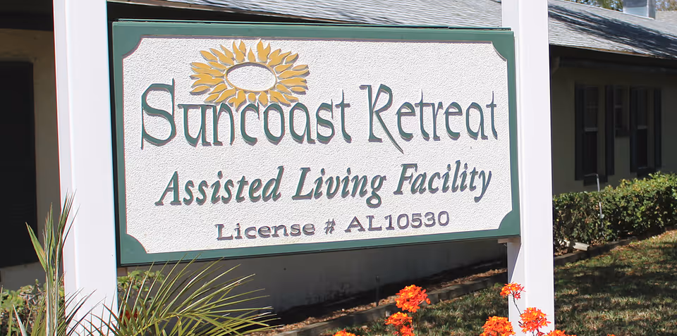 A sign reading "Suncoast Retreat Assisted Living Facility License # AL10530" displayed in front of a building with flowers.