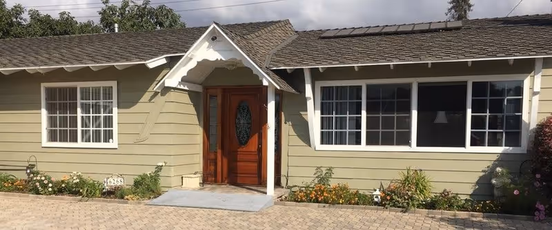 Front exterior view of a single-story building with beige siding, a shingled roof, a wooden front door with an oval glass panel, and two large windows on either side. There are small flower beds with various plants and flowers along the front of the building, and a paved driveway or walkway leading to the entrance.