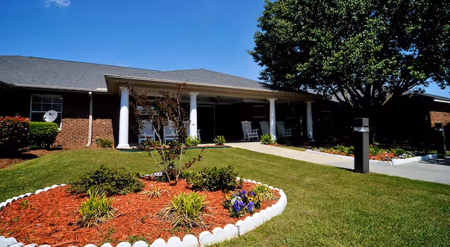 Single-story brick building with a covered porch supported by white columns. The porch has white rocking chairs. In front of the building is a well-maintained lawn with a flower bed bordered by white stones and a large tree on the right side. The sky is clear and blue.
